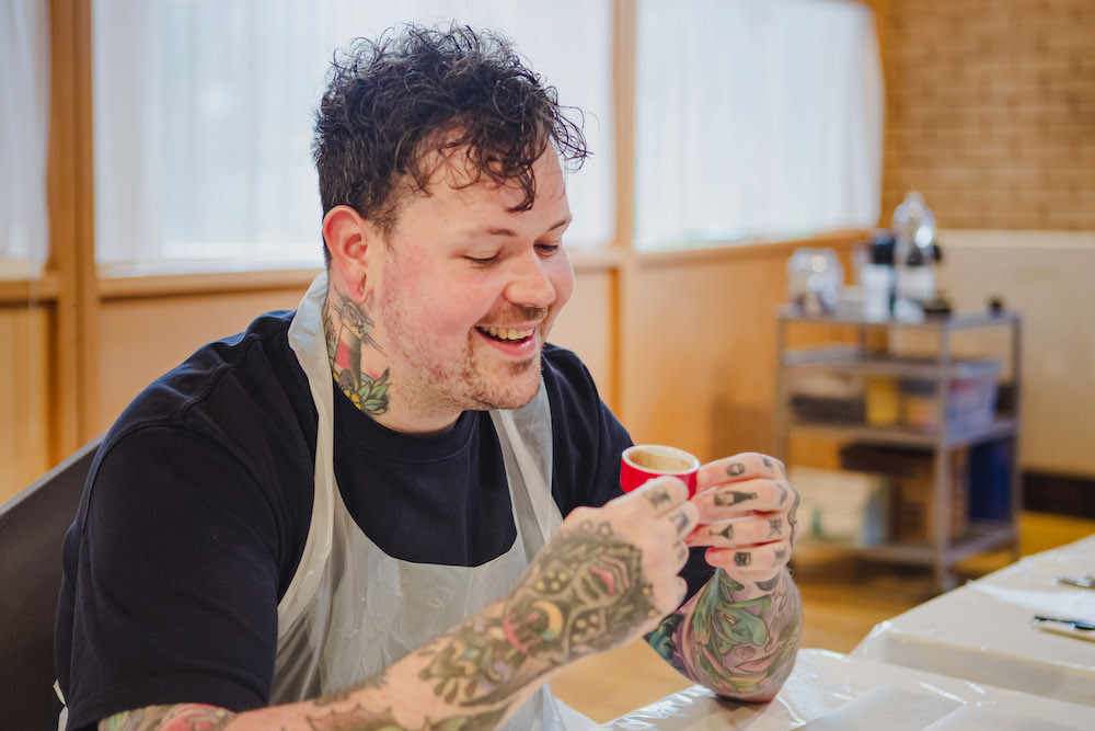 A smiling man with tattooed hands holds a small cup of hot chocolate